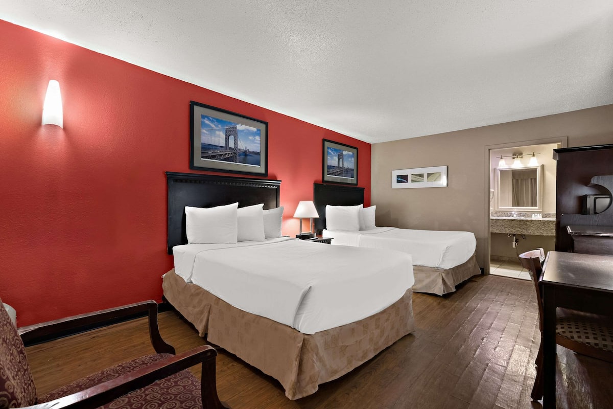 Quarto de hotel com duas camas, piso de madeira, parede de destaque vermelha e banheiro visível por uma porta aberta.