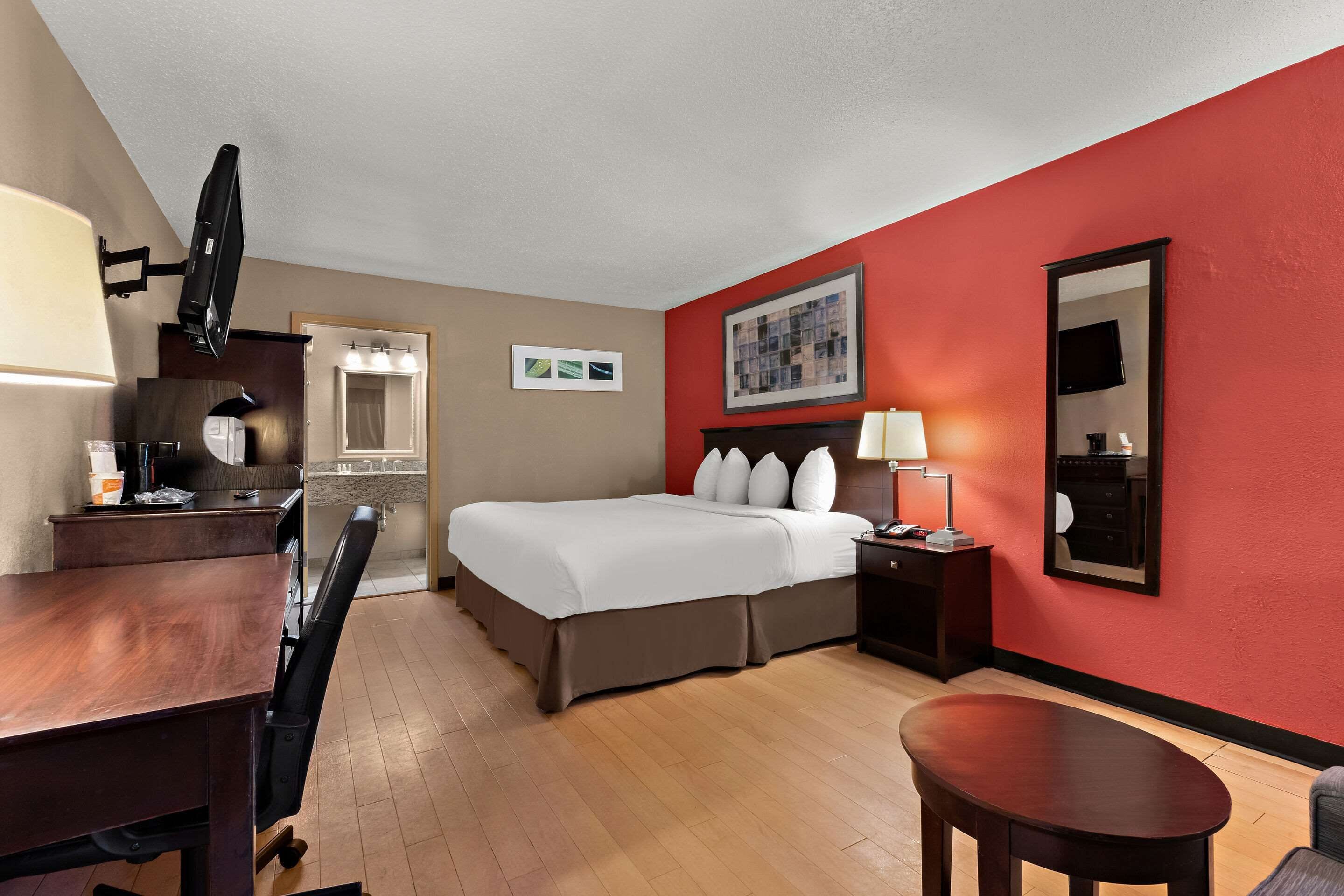 A hotel room with a bed, desk, chair, and a red accent wall.