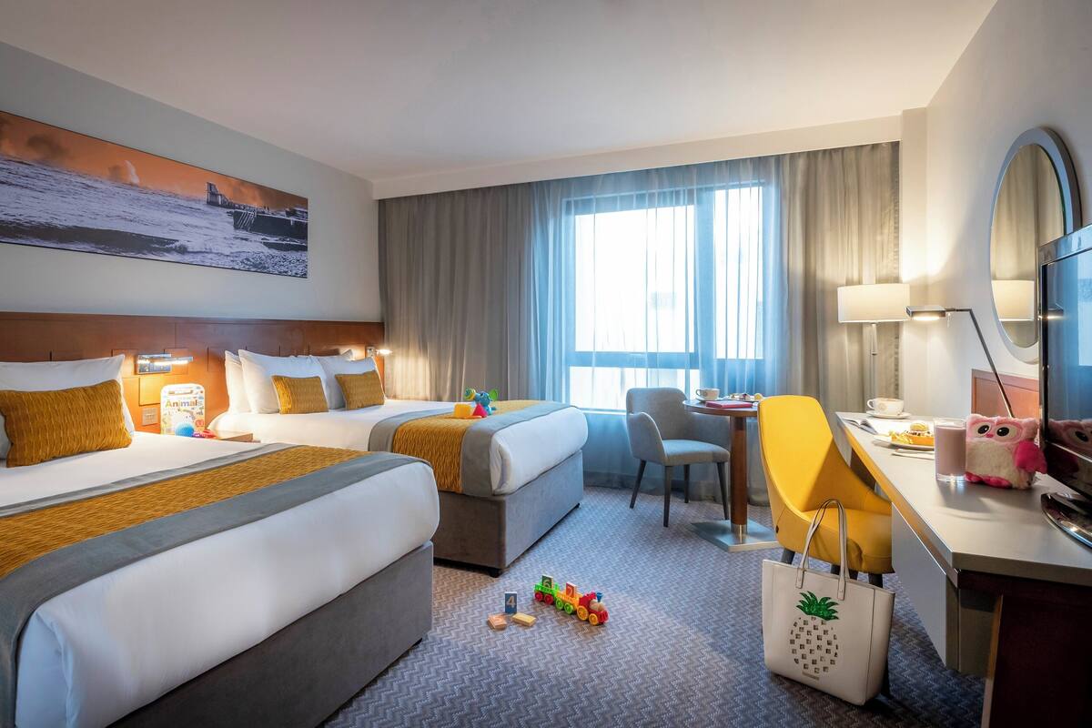 Ein Hotelzimmer mit zwei Betten, einem Schreibtisch, einem Sessel, einem Fernseher und einem großen Fenster mit Vorhängen.