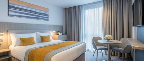 Habitación de hotel con una cama grande, un escritorio y una silla. Se ve una ventana con cortinas, un cuadro en la pared y una pequeña mesa con un jarrón de flores.