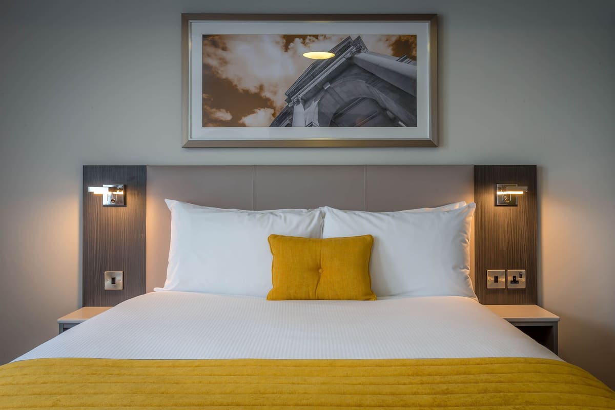 Ein Hotelzimmer mit einem großen Bett, weißer Bettwäsche, einer senffarbenen Tagesdecke und einem gerahmten Bild eines Gebäudes an der Wand.