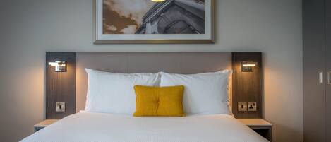A hotel room with a large bed, white bedding, a mustard-colored throw blanket, and a framed picture of a building on the wall.