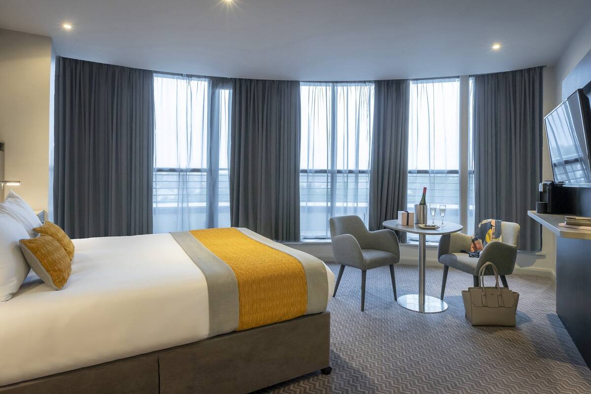Ein Hotelzimmer mit einem Bett, einem Schreibtisch mit Fernseher, einem Sessel, einem kleinen Tisch mit Flasche und Gläsern sowie großen Fenstern mit Vorhängen.