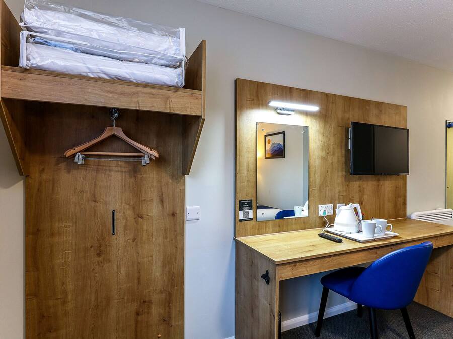 A hotel room with a wooden desk, a blue chair, a bed with a wooden headboard, a hanging coat rack, a flat-screen TV mounted on the wall, and a small table with a kettle and cups.
