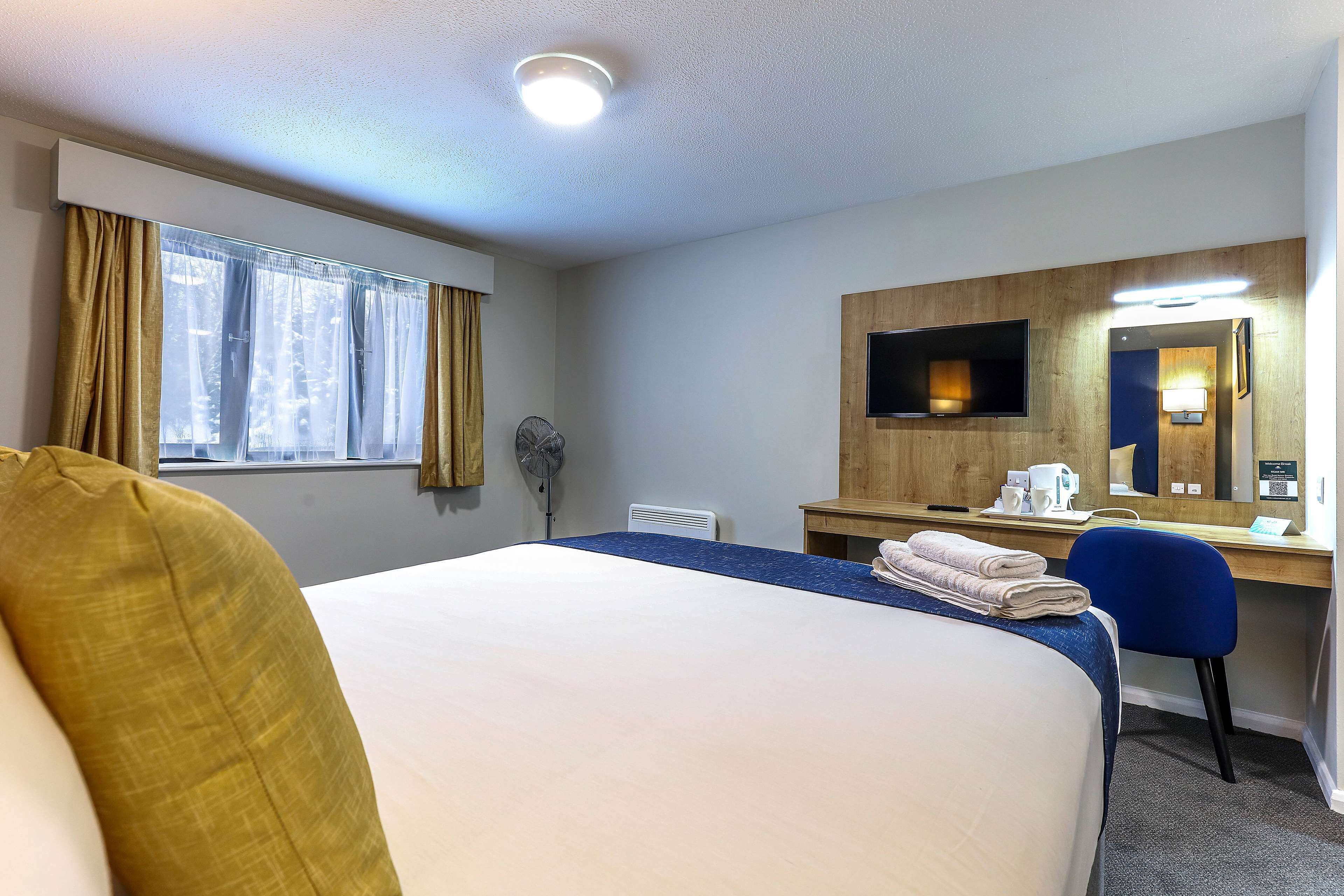 Een hotelkamer met een groot bed, een bureau met een computer en een spiegel, een raam met gordijnen en een plafondlamp.