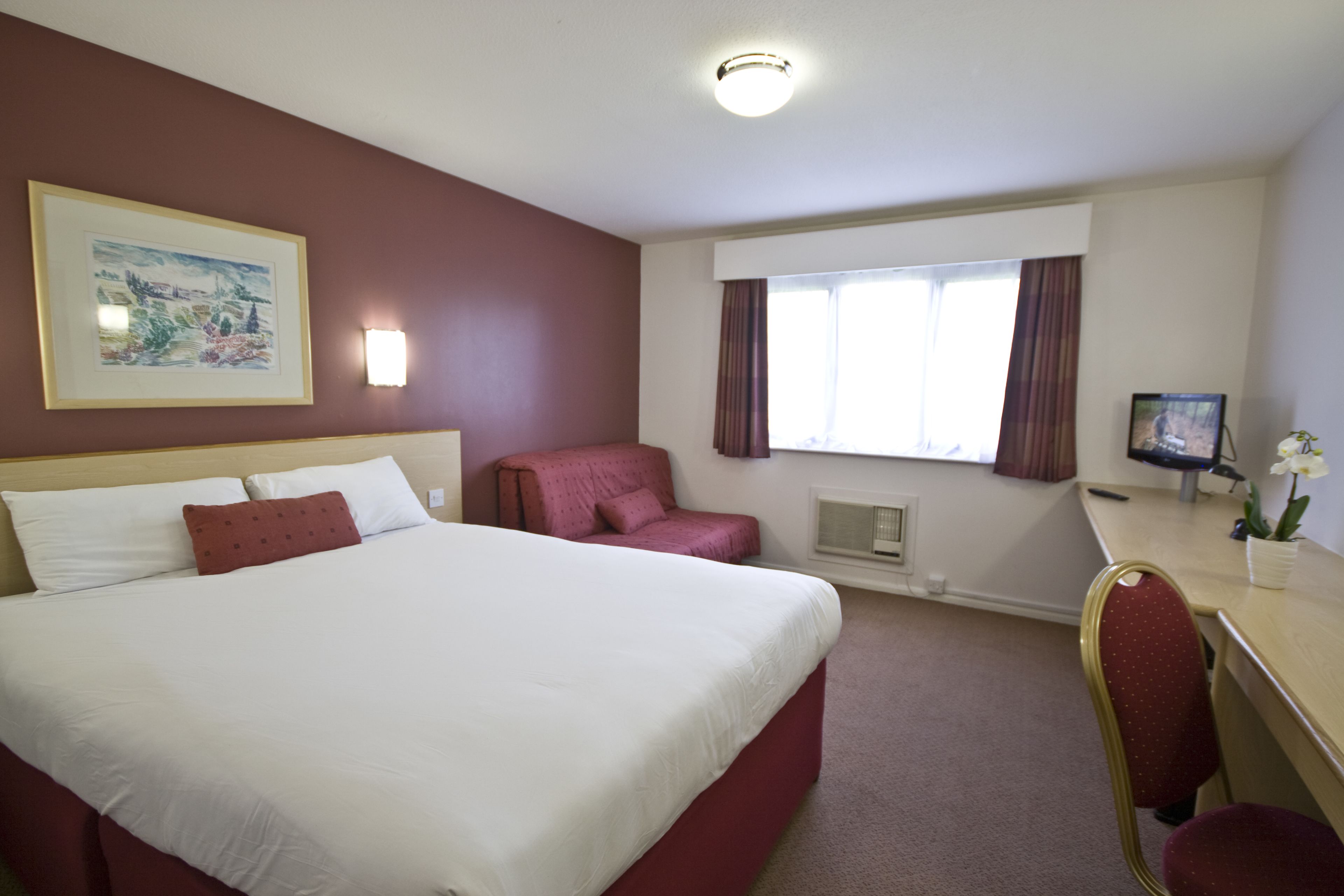 A hotel room with a large bed, a desk, a chair, a window with curtains, and a framed picture on the wall.