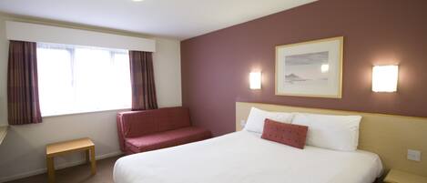 Habitación de hotel con una cama grande, un pequeño banco, un sofá rojo, una ventana con cortinas y luces fijadas a la pared.