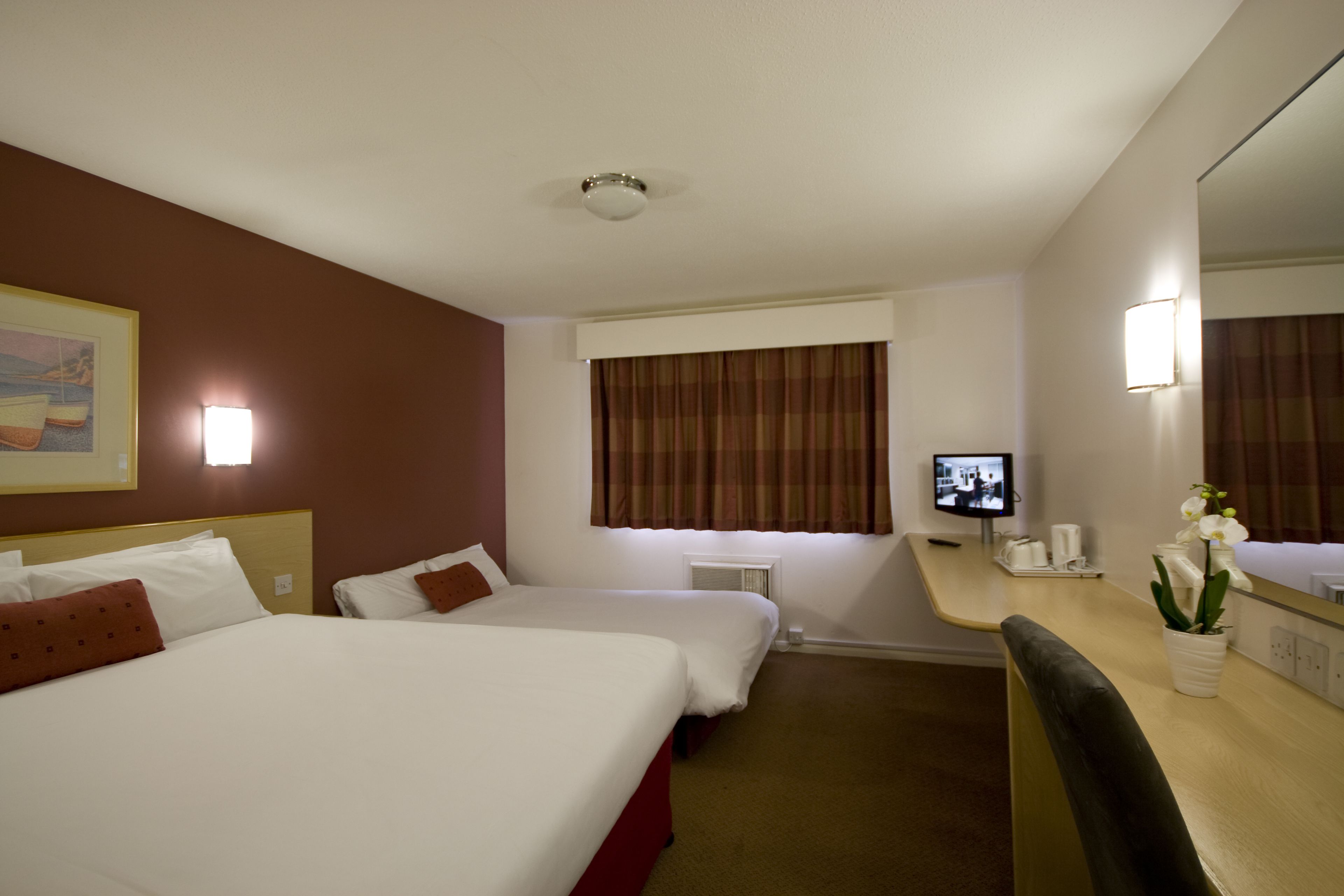 A hotel room with two beds, a desk with a mirror, a television, and a potted orchid.