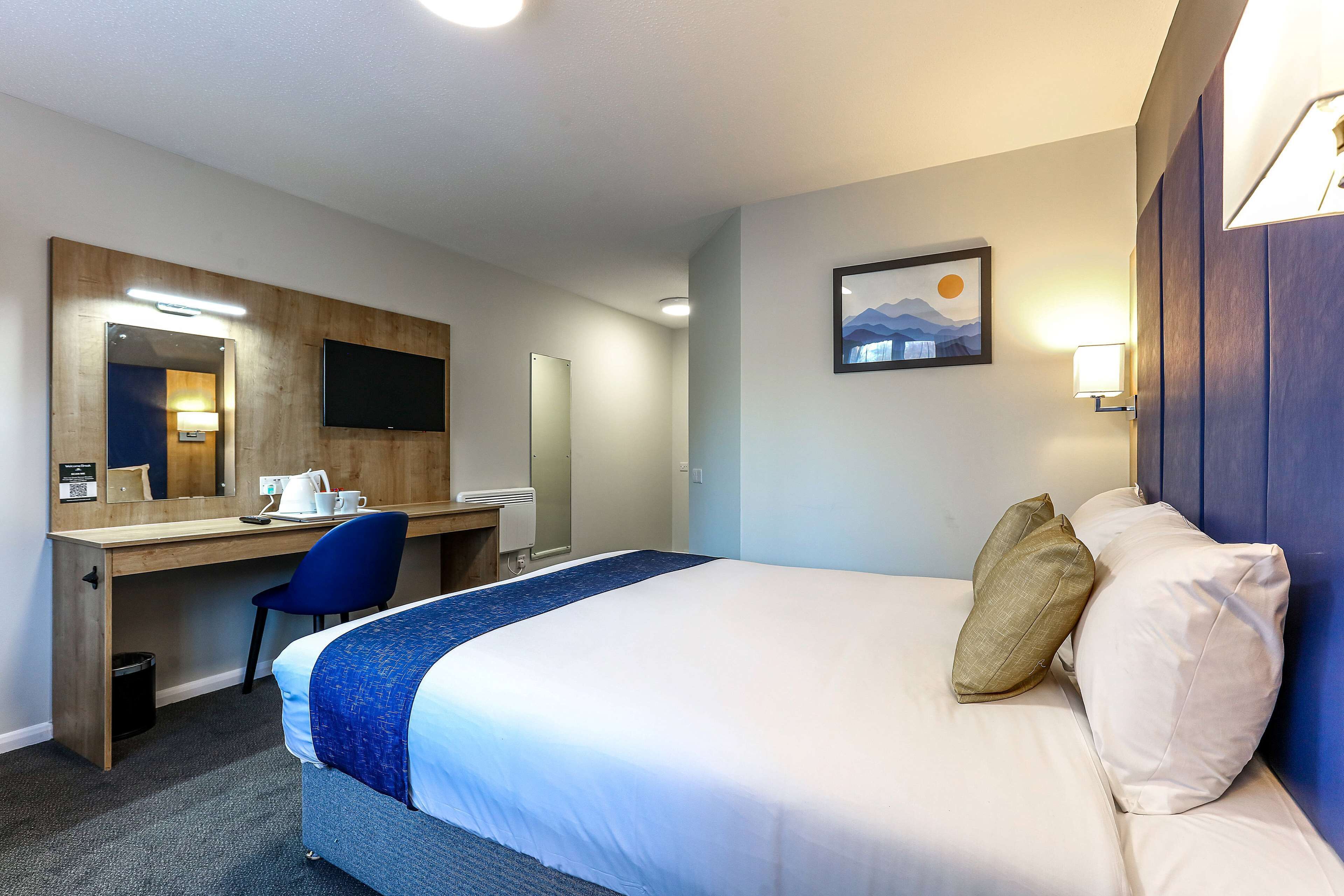 A hotel room with a bed, a desk, a chair, a desk lamp, a trash bin, and a wall-mounted TV.