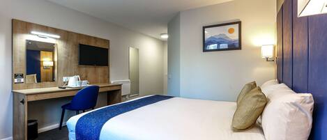 A hotel room with a bed, a desk, a chair, a desk lamp, a trash bin, and a wall-mounted TV.