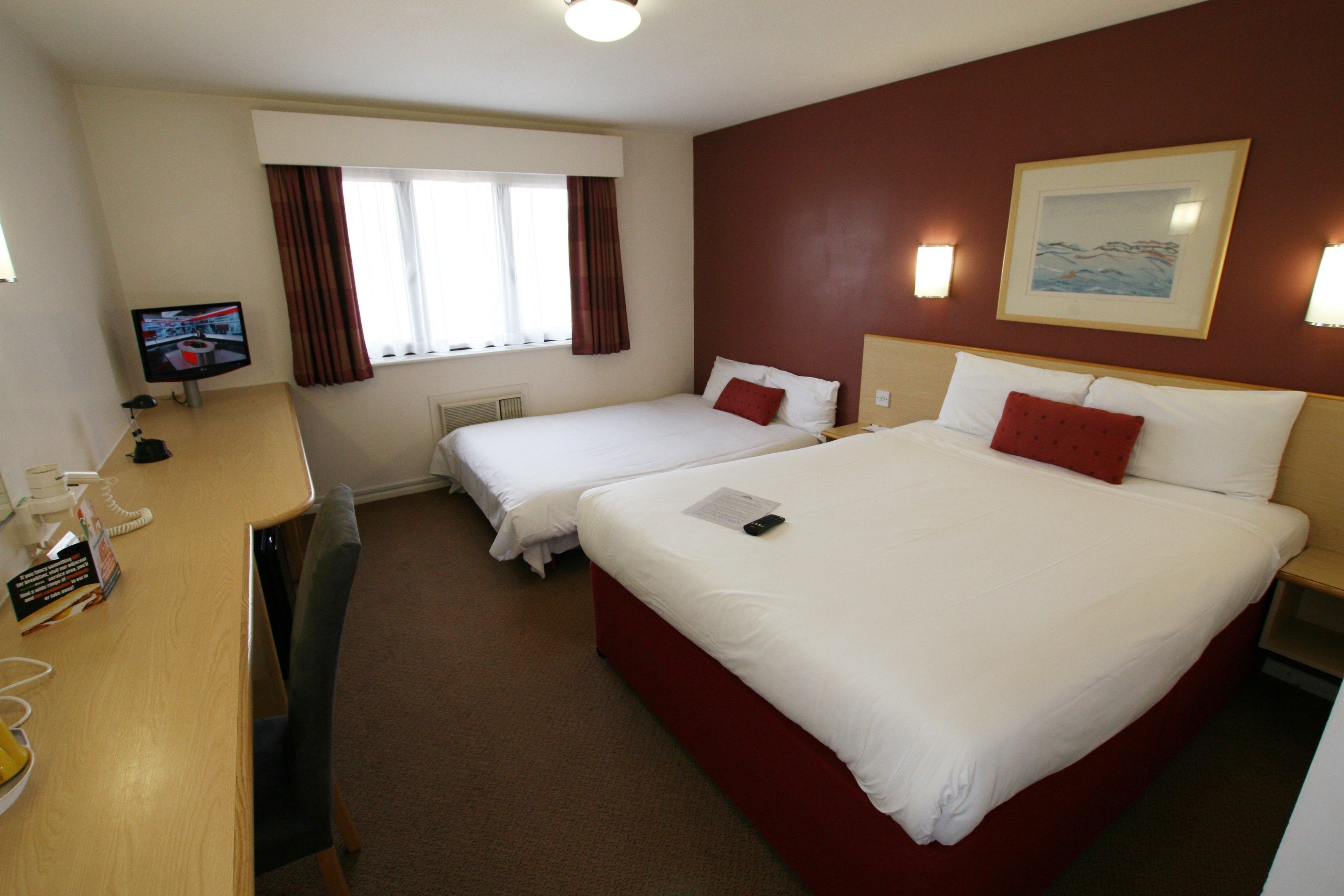 A hotel room with two beds, a desk, a TV, and a window with curtains.