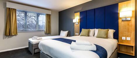 A hotel room with two beds, a large window with curtains, a wall-mounted lamp, and a small wooden shelf.