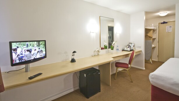 A hotel room with a bed, a desk with a computer, a chair, a TV, and a small fridge. - Days Inn by Wyndham London Stansted Airport (Bishop's Stortford)