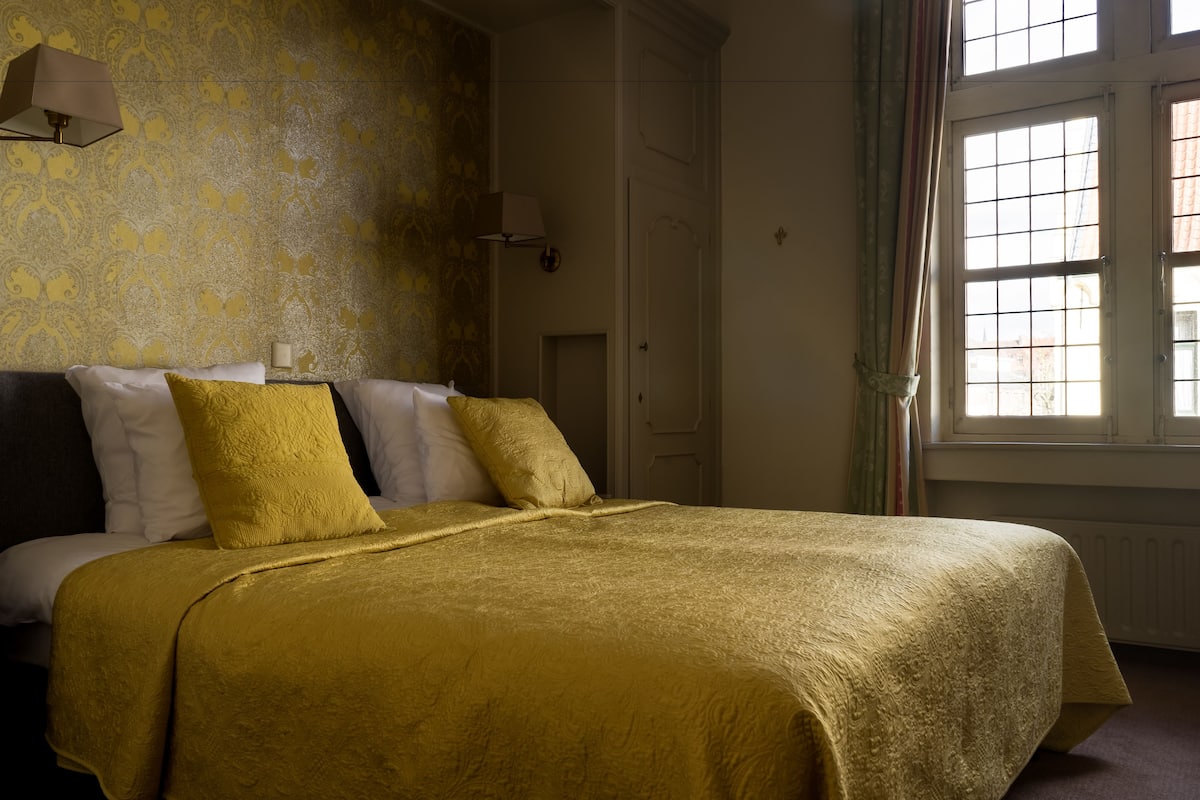 A bed with a gold bedspread and pillows, a nightstand with a lamp, and a window with curtains.