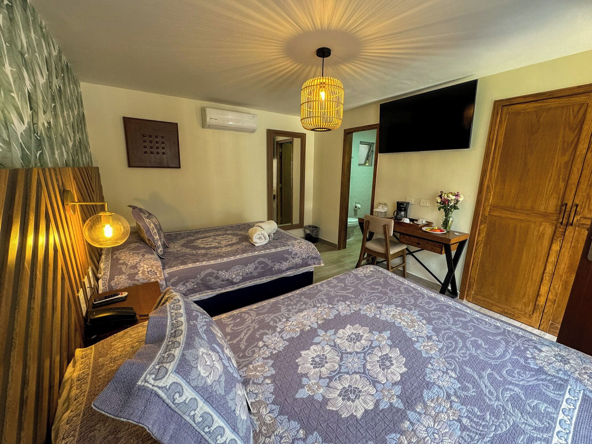 A hotel room with two beds, a desk, a chair, a TV mounted on the wall, and a wooden wardrobe.