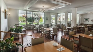 A spacious dining area with multiple tables and chairs, large windows offering views of the outdoors, and a chandelier hanging from the ceiling. - Hotel Isabel (Guadalajara)