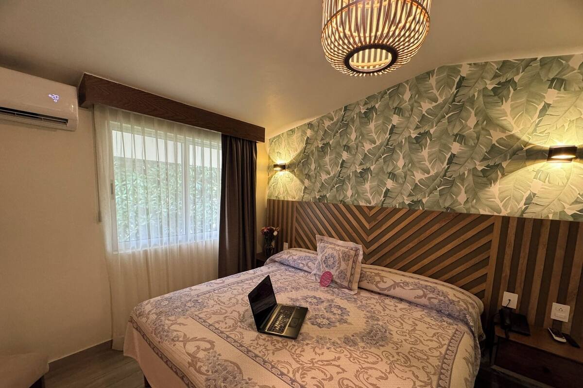 Habitación de hotel con una cama, un ordenador portátil sobre la cama, una lámpara de pared y un foco en el techo.