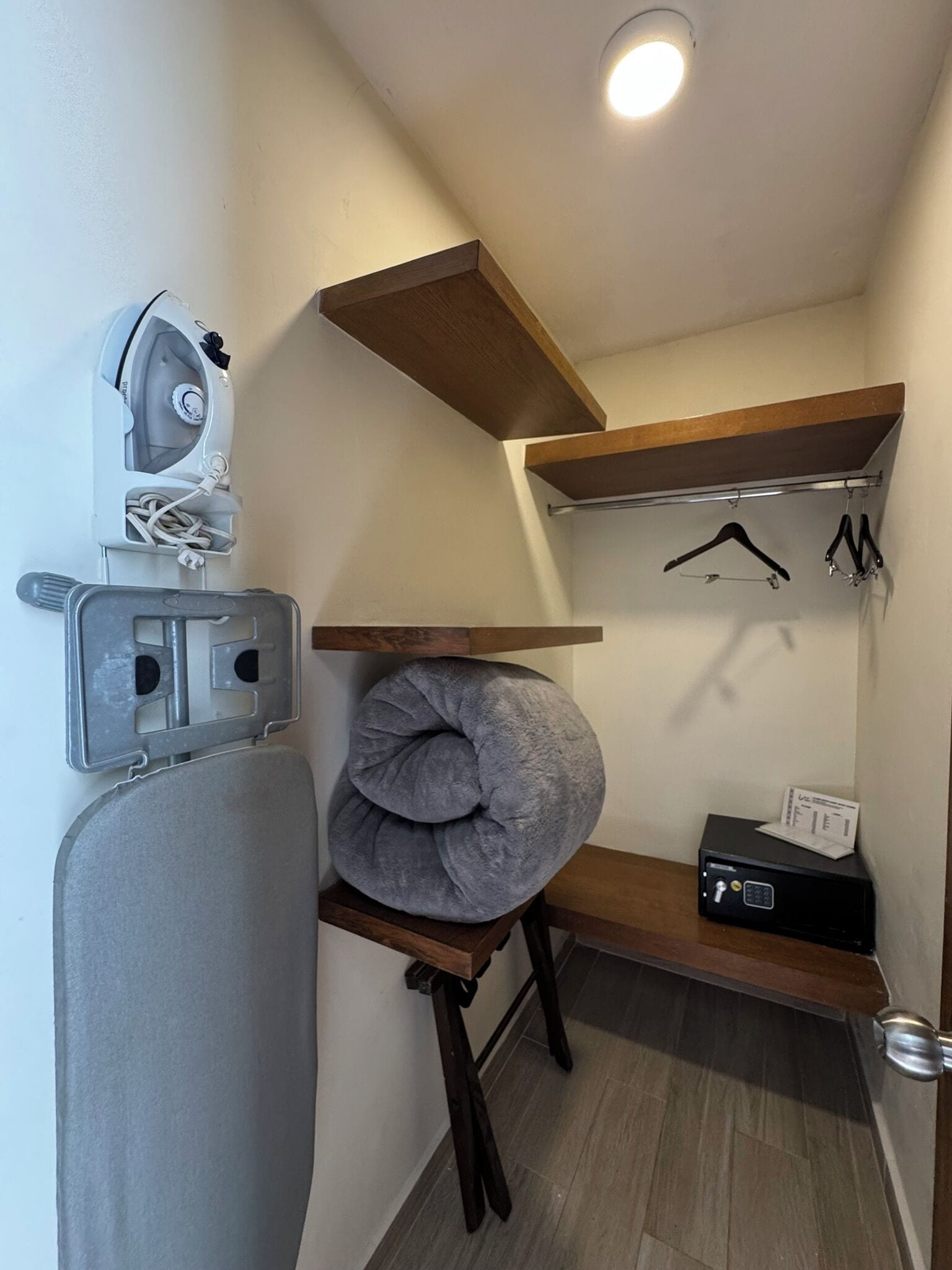 A compact room with a wooden shelf, a gray cushion, a small ironing board, and a black electronic device.