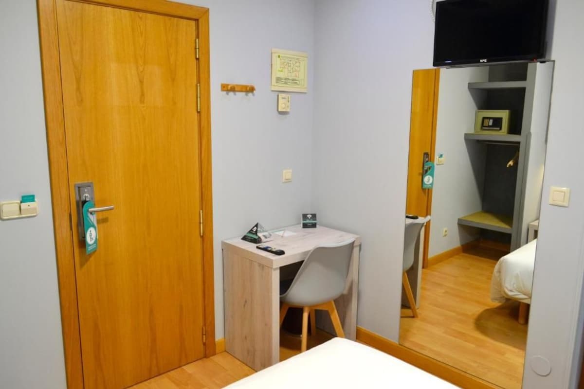 Une petite pièce, de taille modeste, avec une porte en bois, un bureau avec une chaise, un lit et un téléviseur fixé au mur.