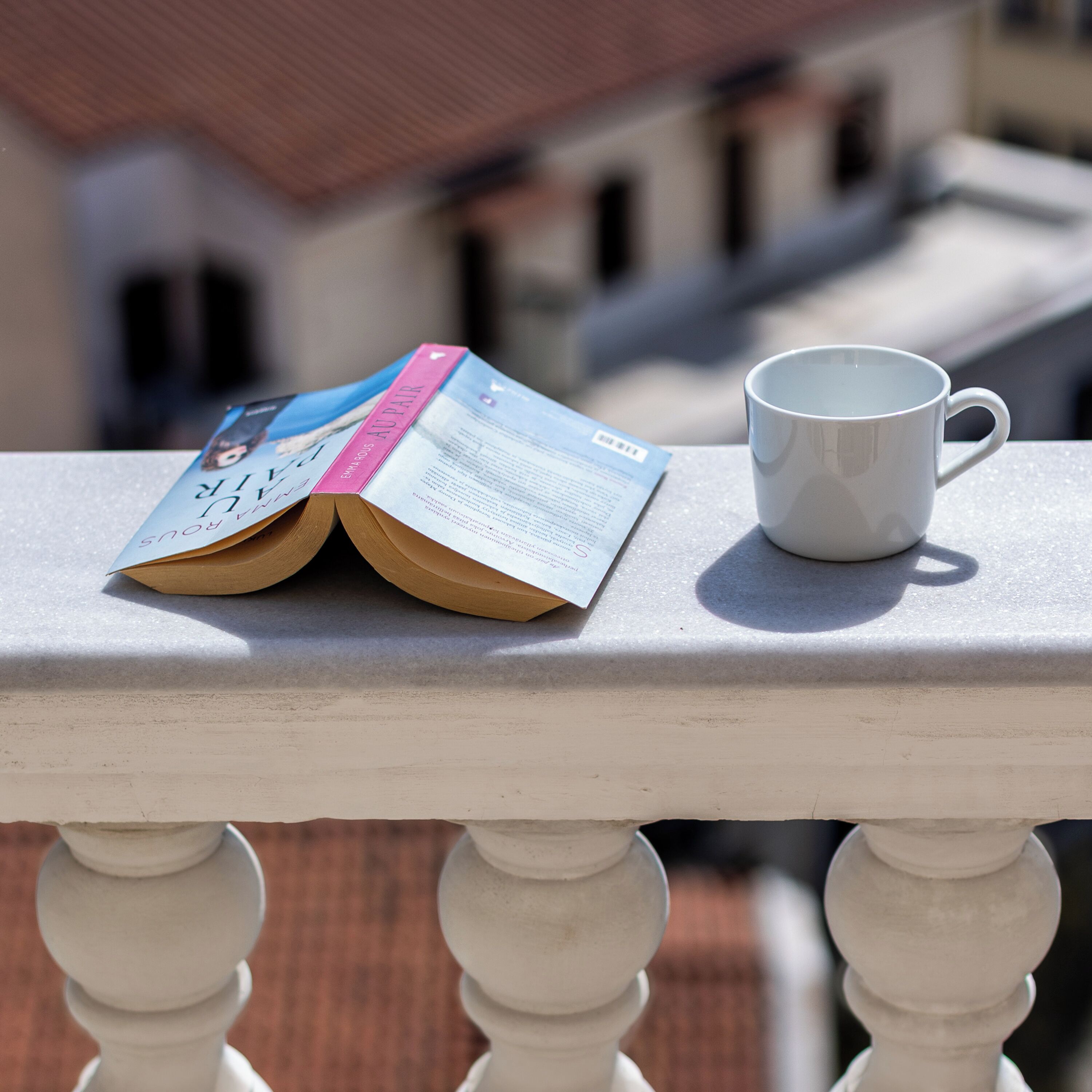 Een boek met de titel 'Au Pair' en een witte mok op een balkonhek.