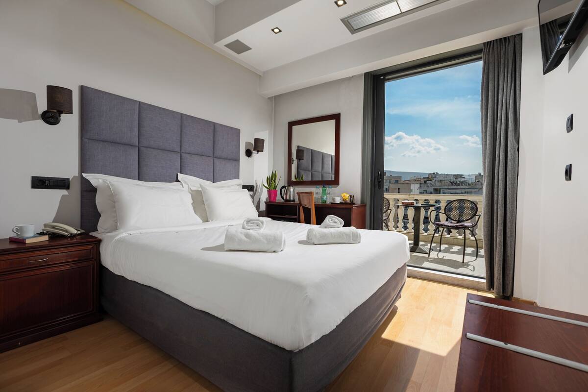 Una habitación de hotel moderna con una cama grande, una mesita de noche con lámpara, una pequeña mesa con silla y un balcón con vistas.