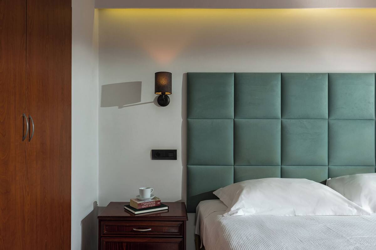 Habitación de hotel con cabecera tapizada en verde, una mesita de noche con lámpara, un pequeño atril y una cama con ropa blanca.