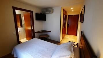 A hotel room with a bed, a desk, a chair, a TV mounted on the wall, and an air conditioning unit.