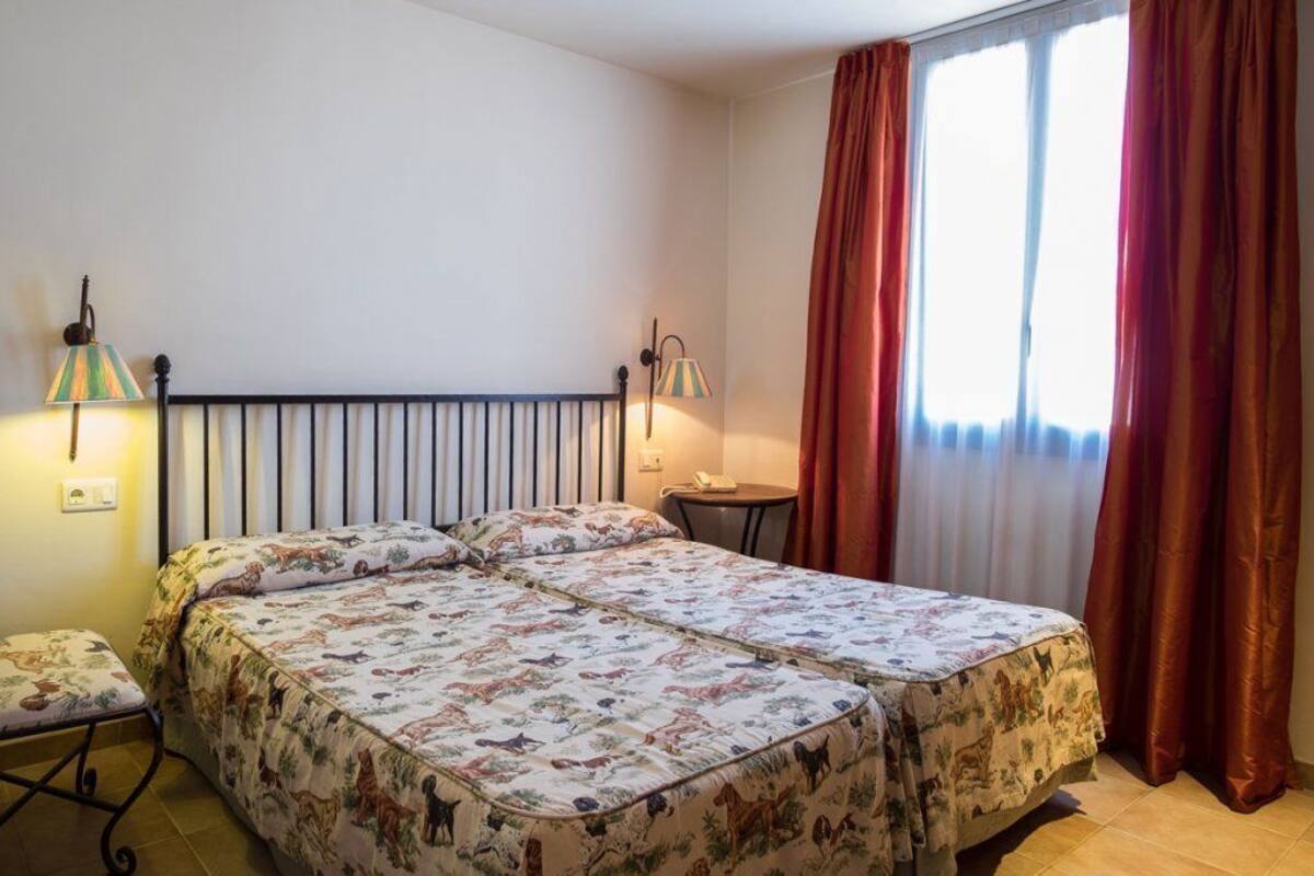 A double bed with a floral bedspread, a small bedside table, a lamp, and a window with curtains.