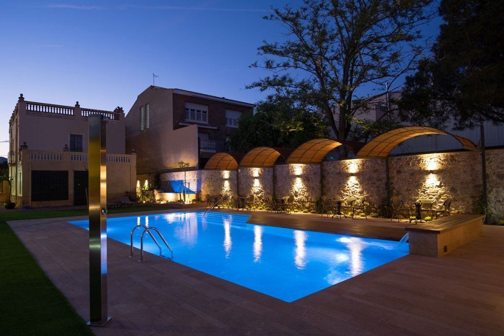 Una piscina con pared de piedra e iluminación en arco, rodeada por una terraza de madera y un edificio al fondo.