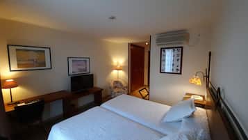A hotel room with a bed, a desk with a TV, a chair, a lamp, and a wall-mounted air conditioner.