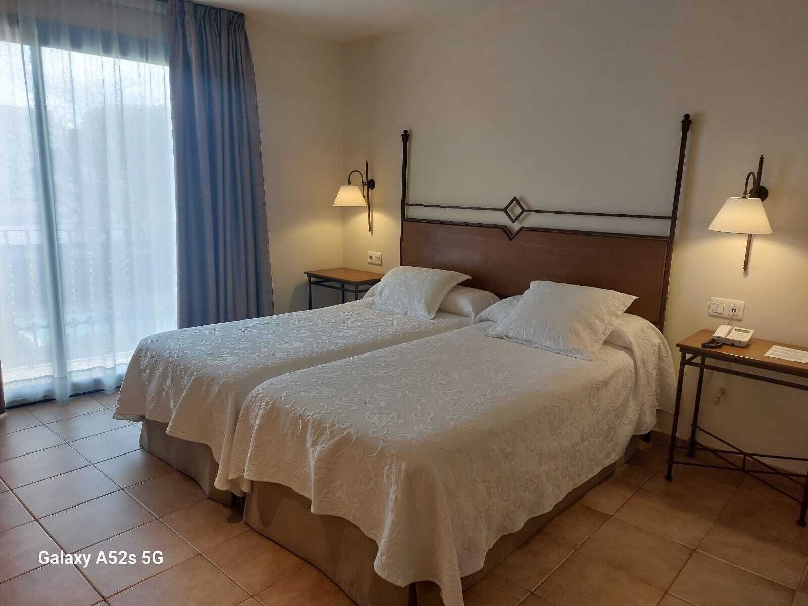 Hotellihuone, jossa on kaksi sänkyä, yöpöytä lampun kanssa ja ikkuna, jossa on verhot.