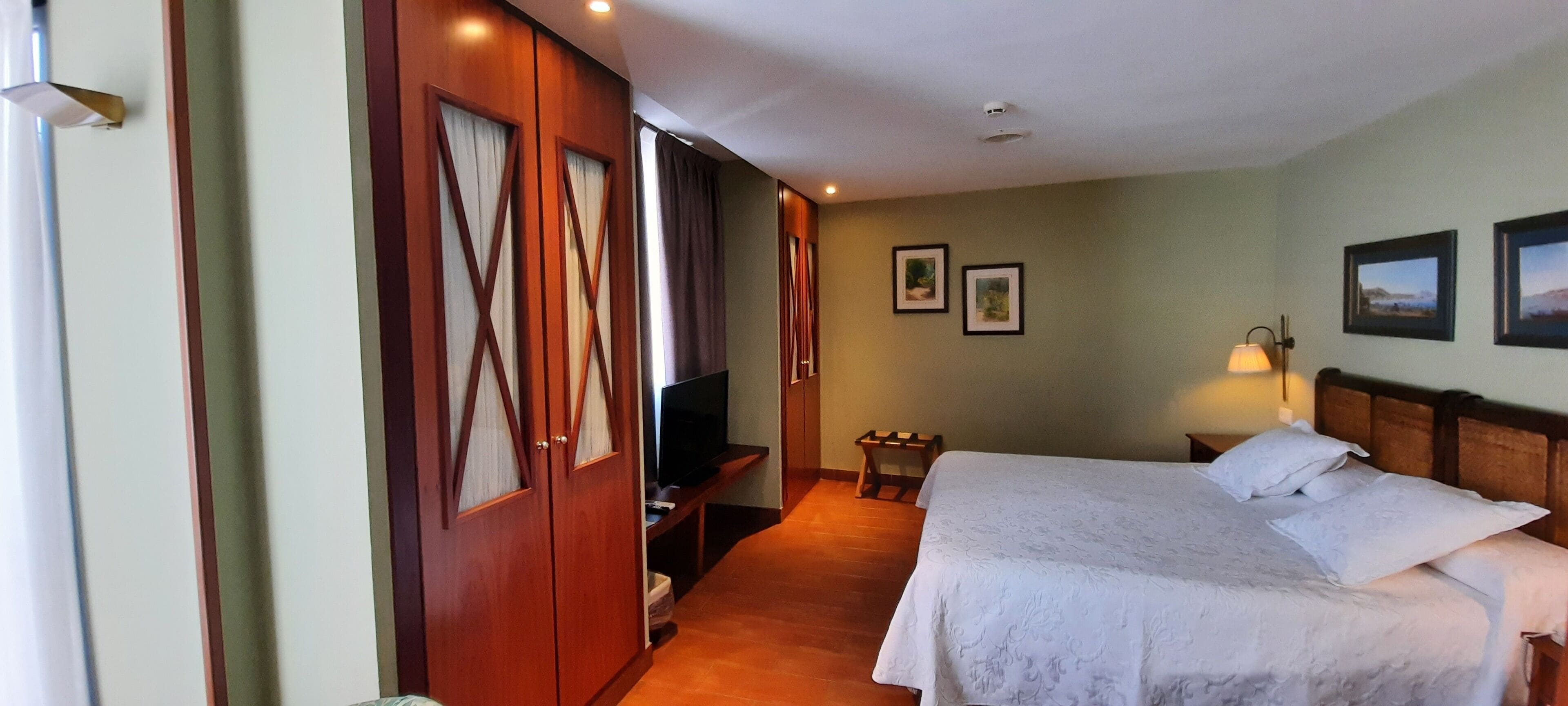 Una habitación de hotel con una cama, televisión, armario de madera y una mesita con lámpara.