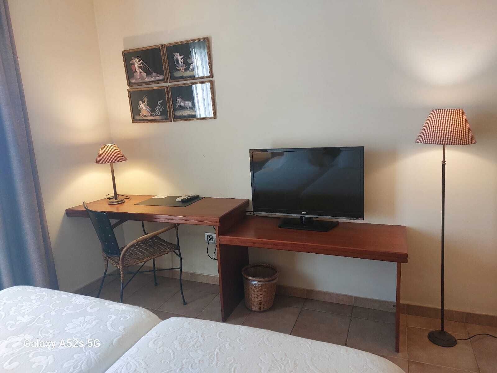 A hotel room with a wooden desk, a television, a chair, a bed, and a lamp.