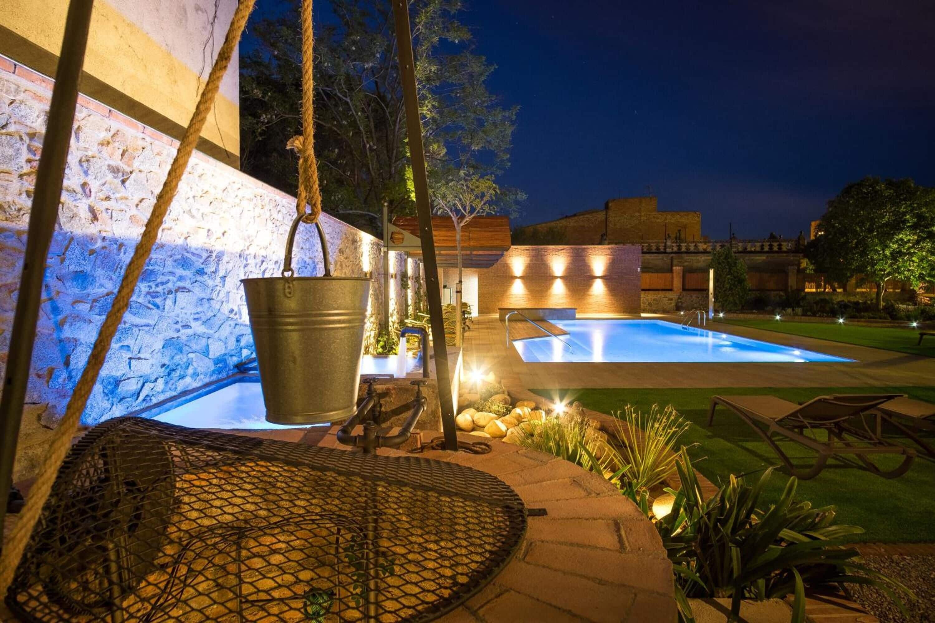 A swimming pool area with a stone wall, a hanging metal bucket, and a lounge chair.
