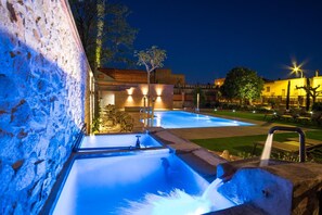 Zona de piscina exterior de noche con pared de piedra, fuente de agua y sillones de descanso.