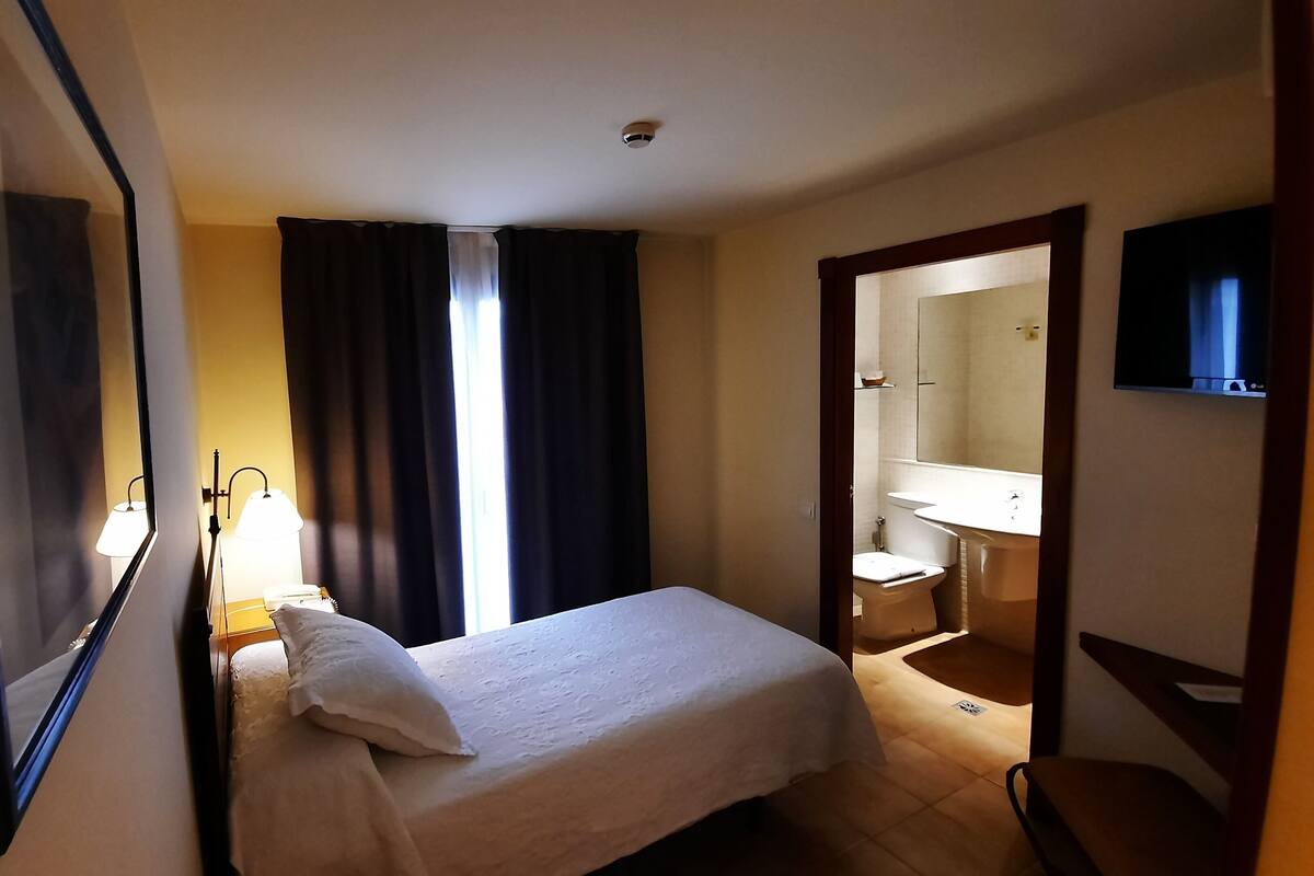 A hotel room with a bed, a nightstand, a lamp, a window with curtains, a bathroom with a sink and mirror, and a television.