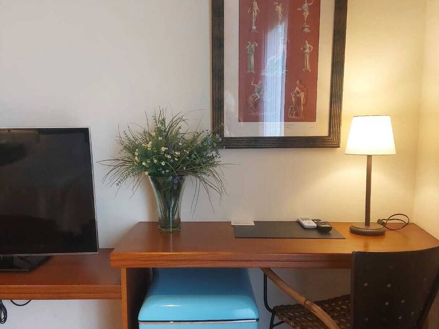 A room with a TV, a desk with a lamp, a chair, a small blue cabinet, and a framed picture on the wall.
