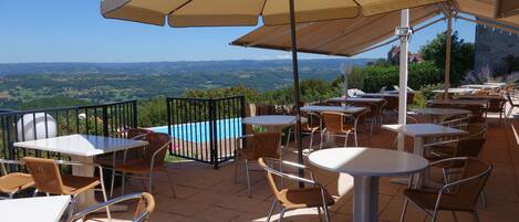 Um pátio externo com mesas, cadeiras, piscina e uma vista panorâmica.