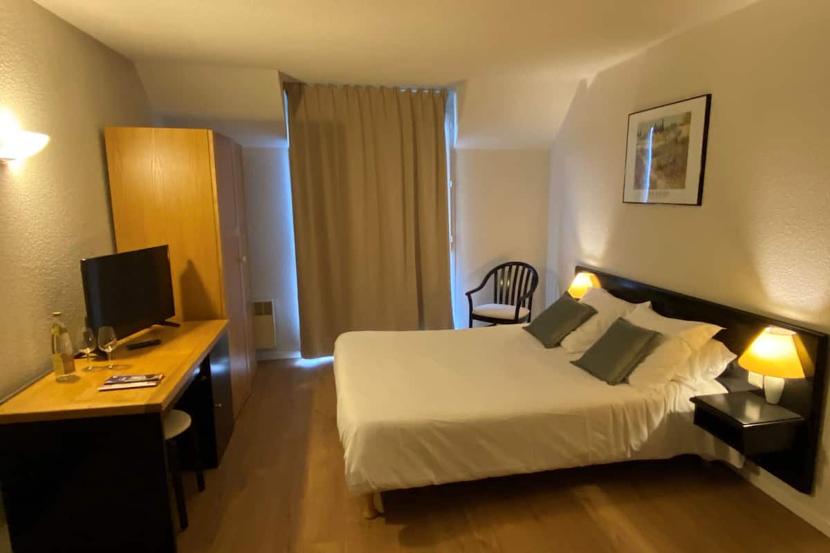 A hotel room with a bed, bedside tables, a desk with a computer, a chair, and a window with curtains.