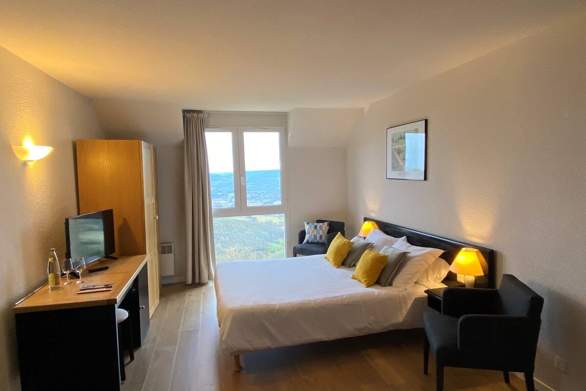 A hotel room with a bed, a desk with a computer, a chair, and a window with a view of the countryside.
