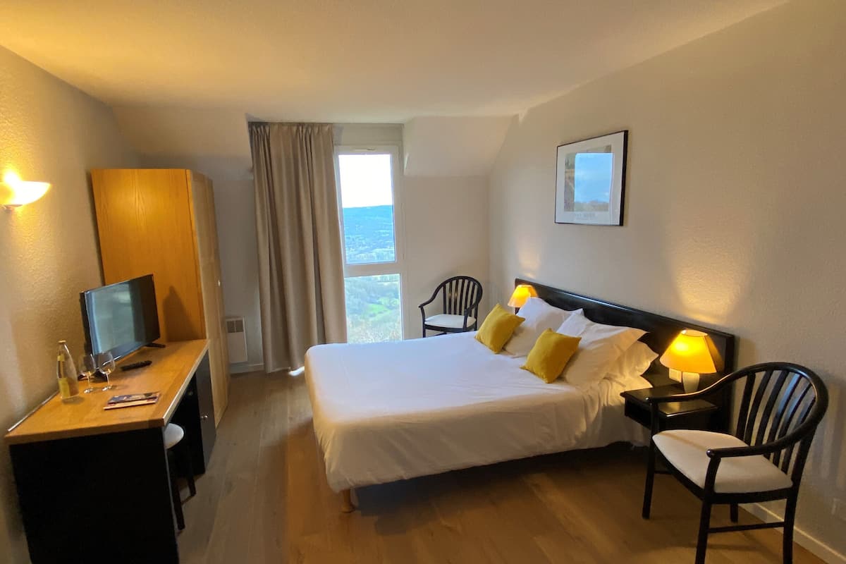A hotel room with a bed, bedside tables, a desk with a computer, a chair, and a window with curtains.