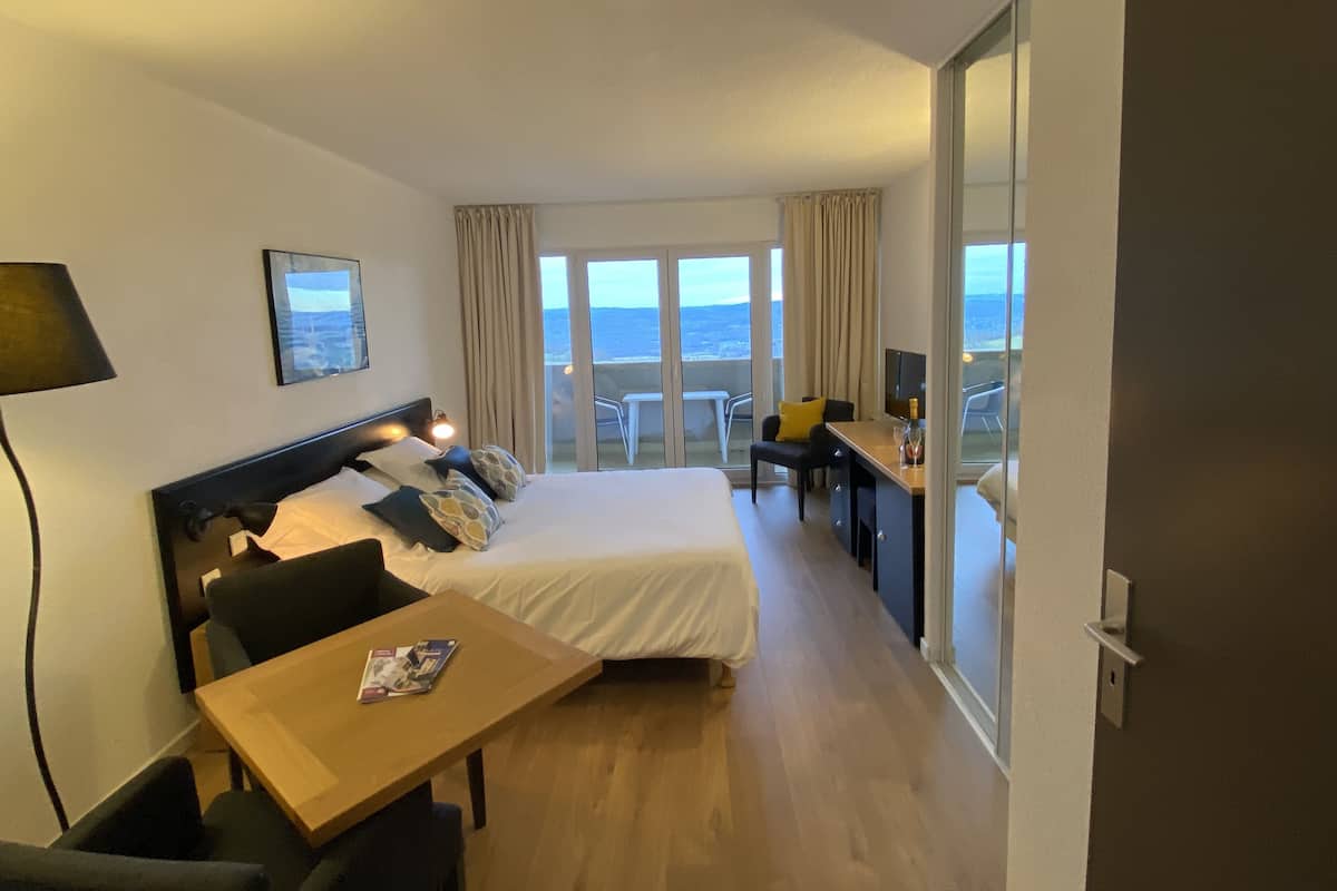 A hotel room with a bed, a desk, a chair, and a view of the sea through large windows.