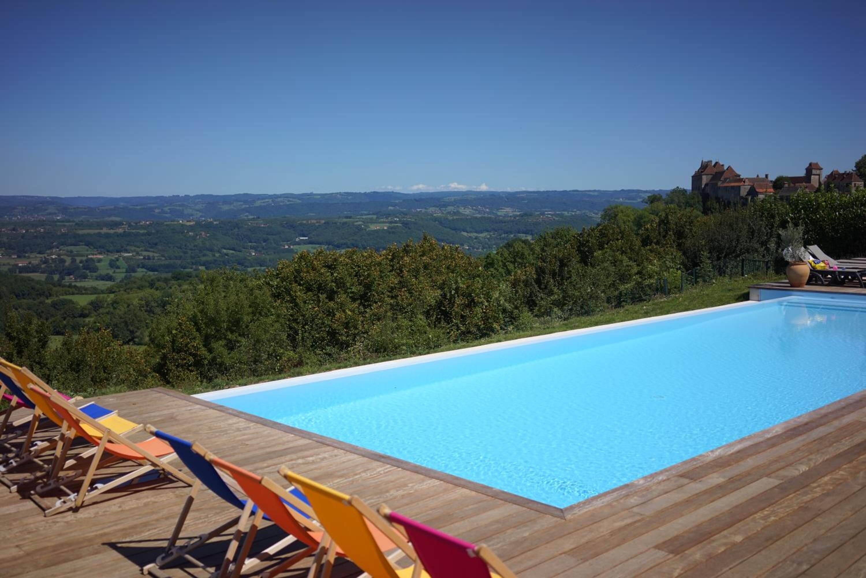 A swimming pool with wooden deck and lounge chairs, surrounded by greenery and a view of distant hills.