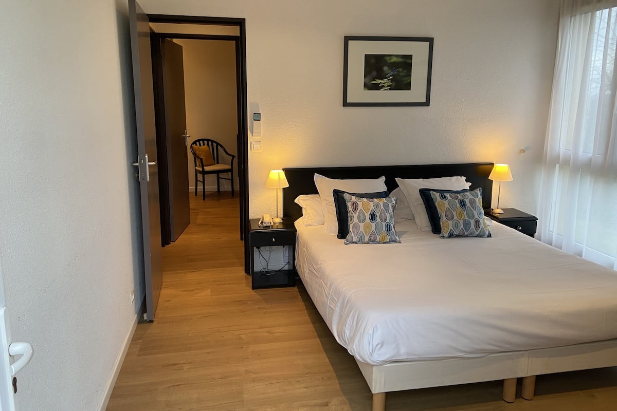 A hotel room with a large bed, two bedside tables with lamps, a framed picture on the wall, and a chair in the adjacent room.