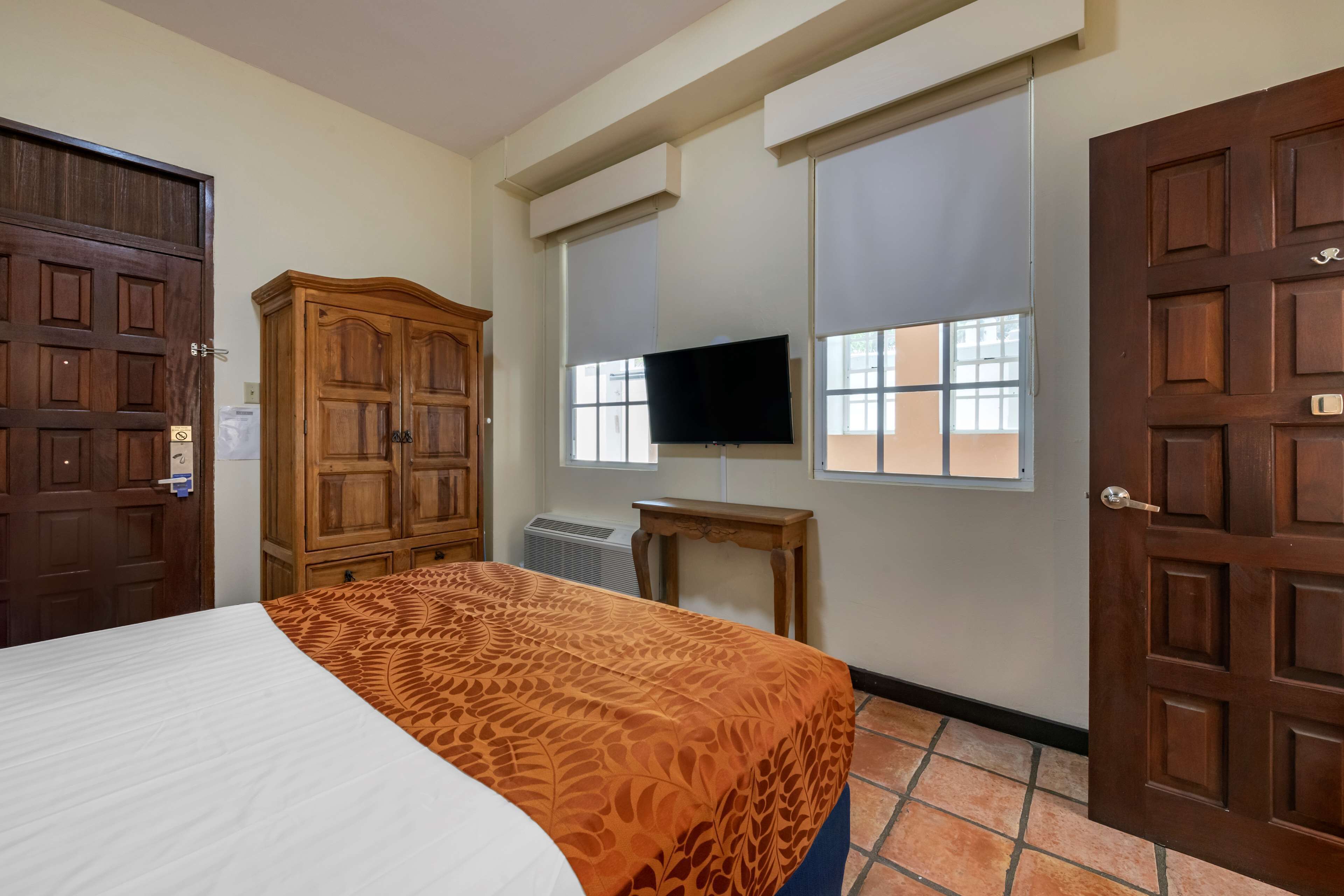 Une chambre d’hôtel avec un lit, une armoire en bois, une télévision et une fenêtre avec des stores.