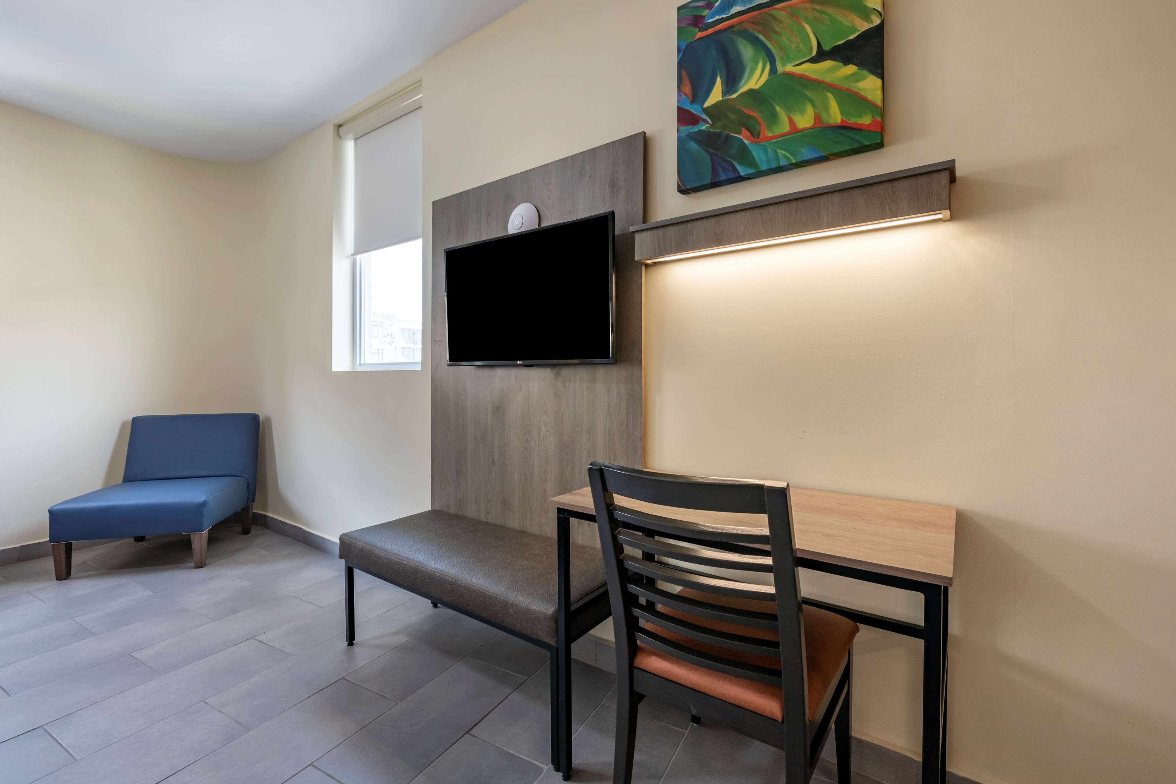 Una habitación moderna con una televisión de pantalla plana montada en un panel de madera, un banco, una silla y un cuadro abstracto de colores en la pared.