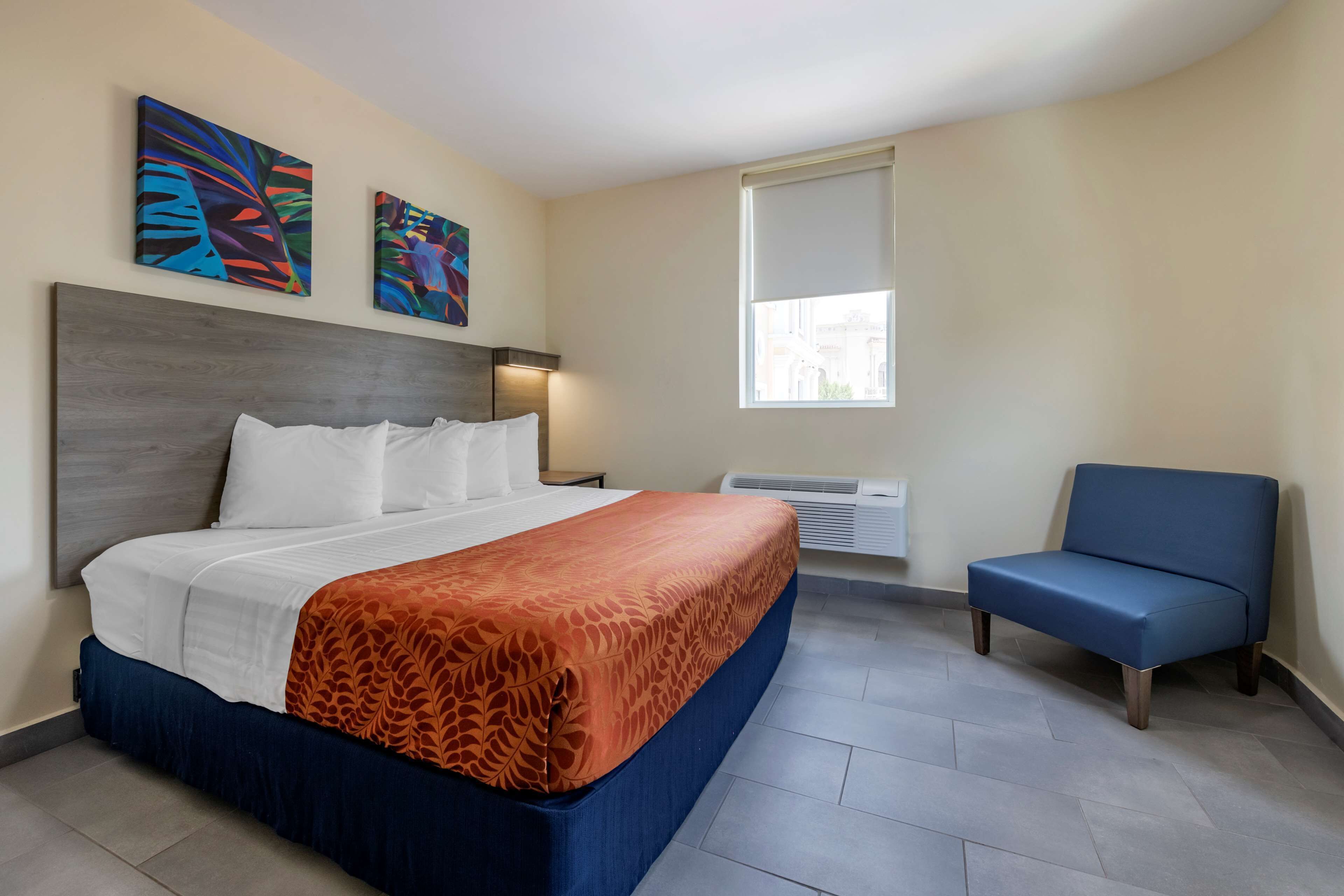 A hotel room with a bed, a blue armchair, and two abstract paintings on the wall.
