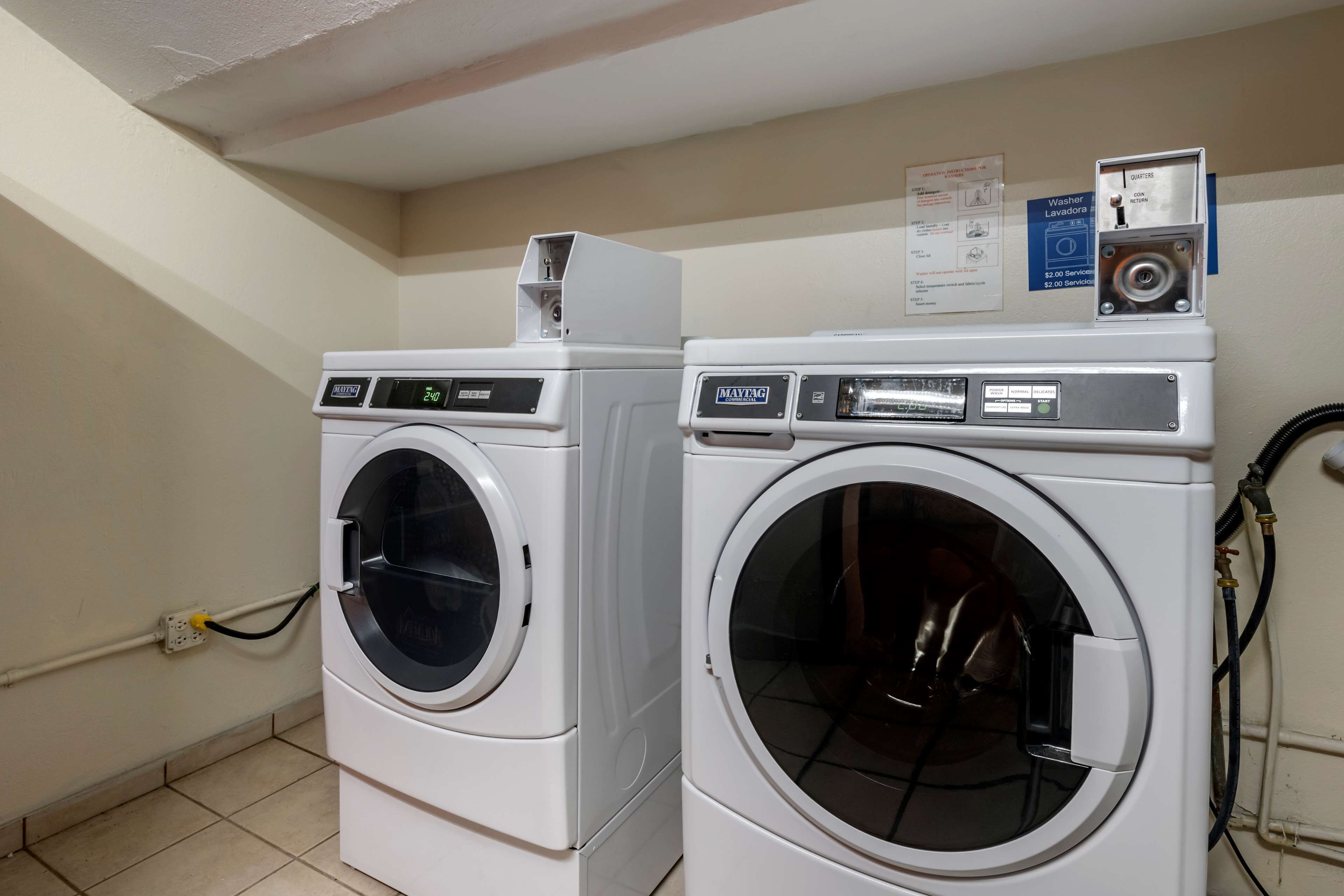 A laundry area with two front-loading washing machines, a wall-mounted paper dispenser, and a sign indicating a $2.00 service fee for washing.