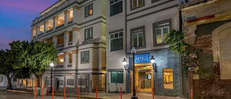 A multi-story hotel with a modern facade, a visible entrance with a sign, and a tree-lined street.