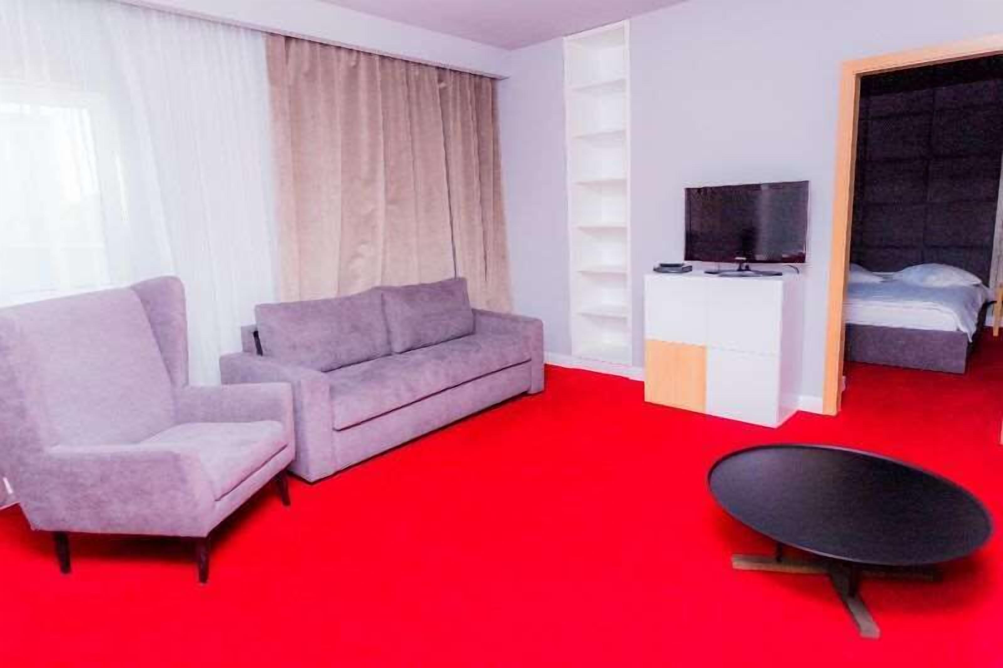 Habitación de hotel con alfombra roja, sofá gris, una mesita negra pequeña, un televisor sobre un mueble blanco y una cama con cabecero oscuro.